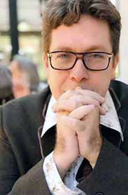 Photo of Mark Malatesta, former literary agent turned author coach and consultant, wearing brown and gold rectangular glasses, a brown suit, and a floral shirt